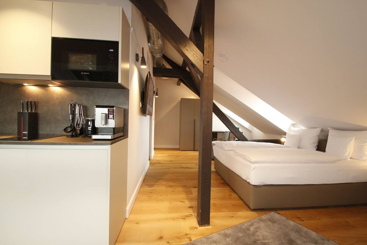 A modern bedroom with a large bed, a kitchenette with a Bosch microwave, and a wooden ceiling beam.