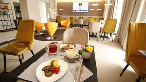A hotel dining area with yellow chairs, a table set with food, a TV screen, and a modern interior design. - Amelie N°1 Hotel & Appartements (Landau in der Pfalz)