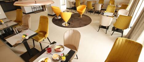 A modern hotel dining area with round tables, yellow chairs, and a TV screen in the background.