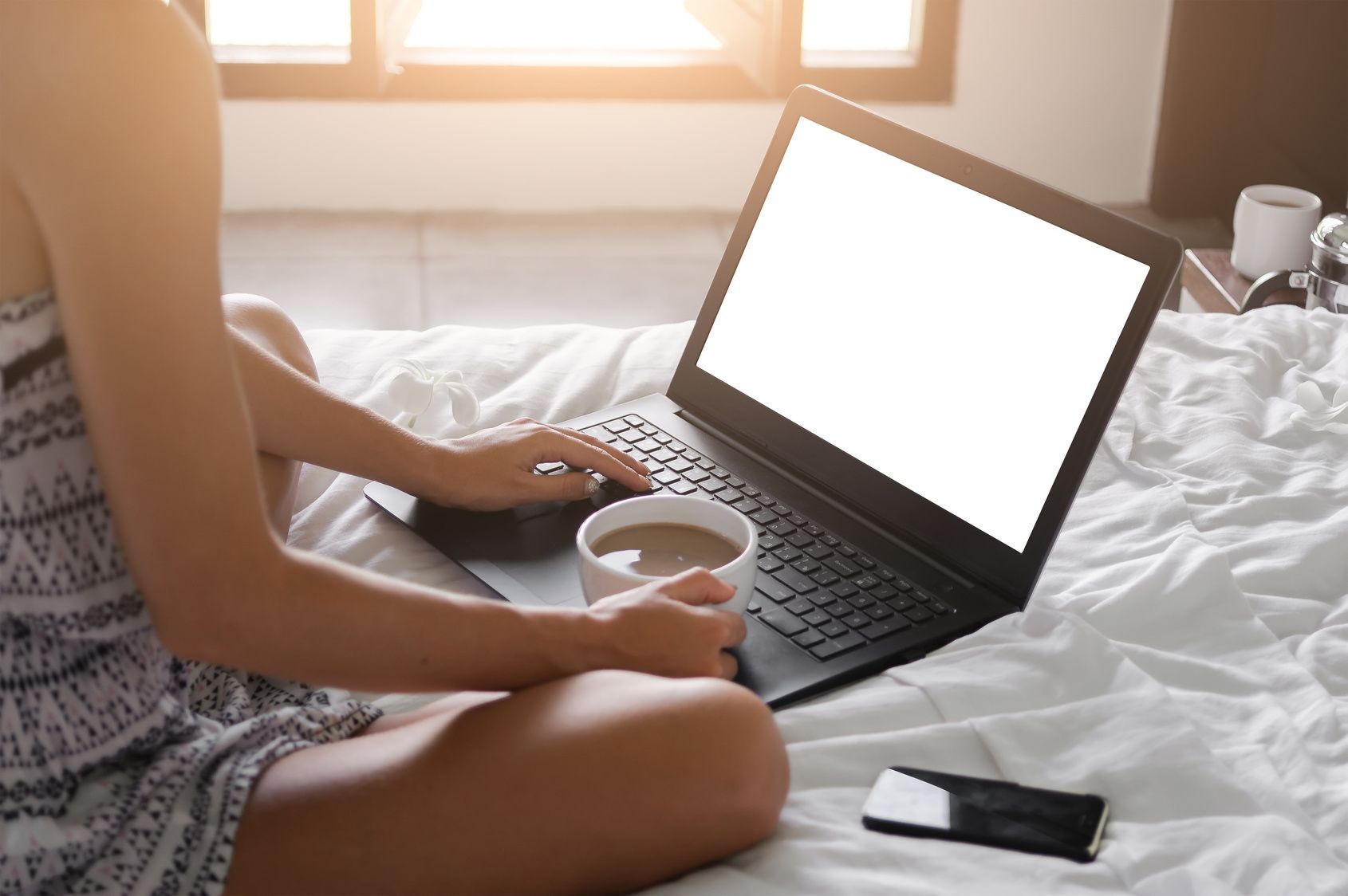 Une personne assise sur un lit, avec un ordinateur portable et une tasse de café.