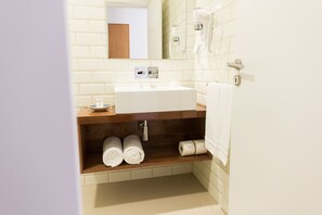 A modern bathroom with a white sink, wooden shelf, and neatly rolled towels. - Apartamentos Praça Amarela (Funchal)