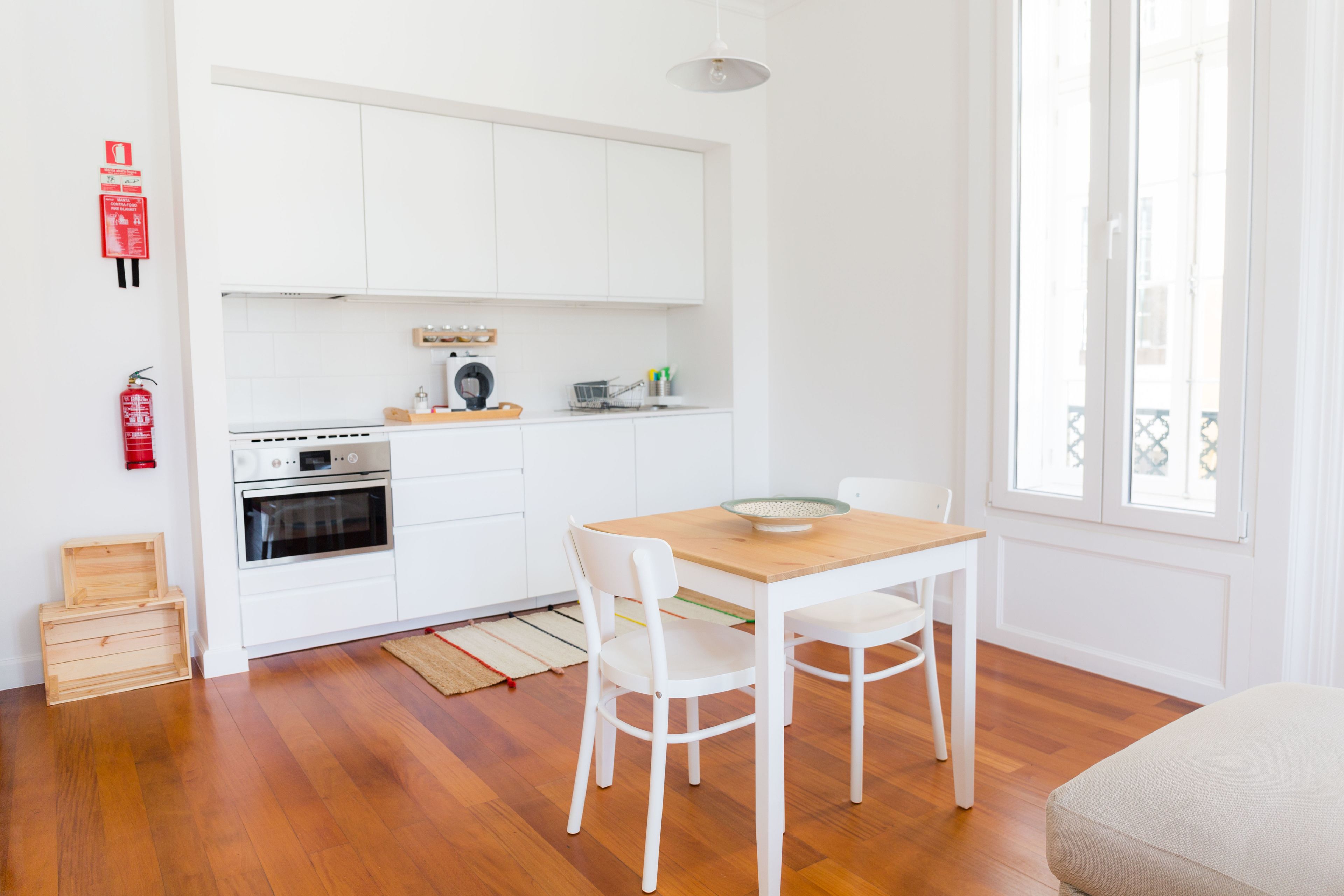 A modern kitchen with white cabinets, a wooden dining table, and chairs. There is a fire extinguisher on the wall and a window with a view of buildings.