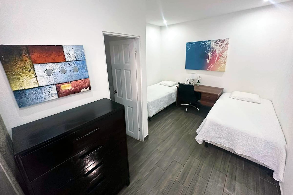 A hotel room with two single beds, a wooden dresser, a desk with a chair, and two wall-mounted paintings.
