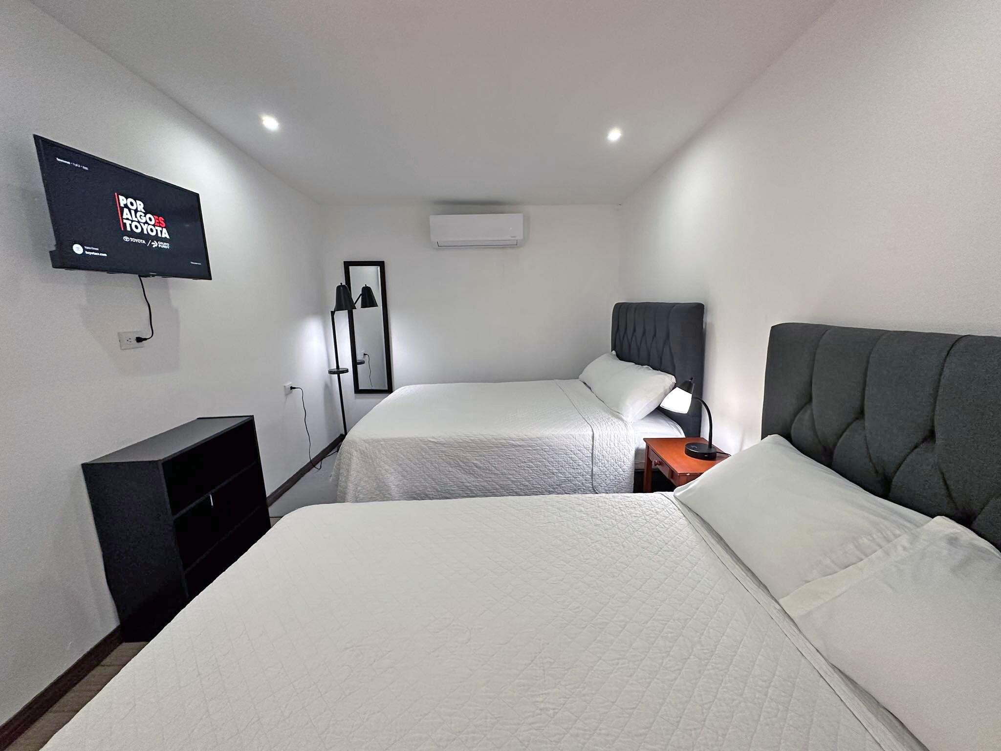 Une chambre d’hôtel avec deux lits, un téléviseur fixé au mur, un miroir et une table de chevet avec une lampe.