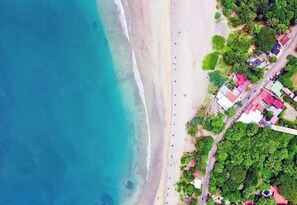 An aerial view of a beach with a road running parallel to the shore, lined with colorful buildings and lush greenery.