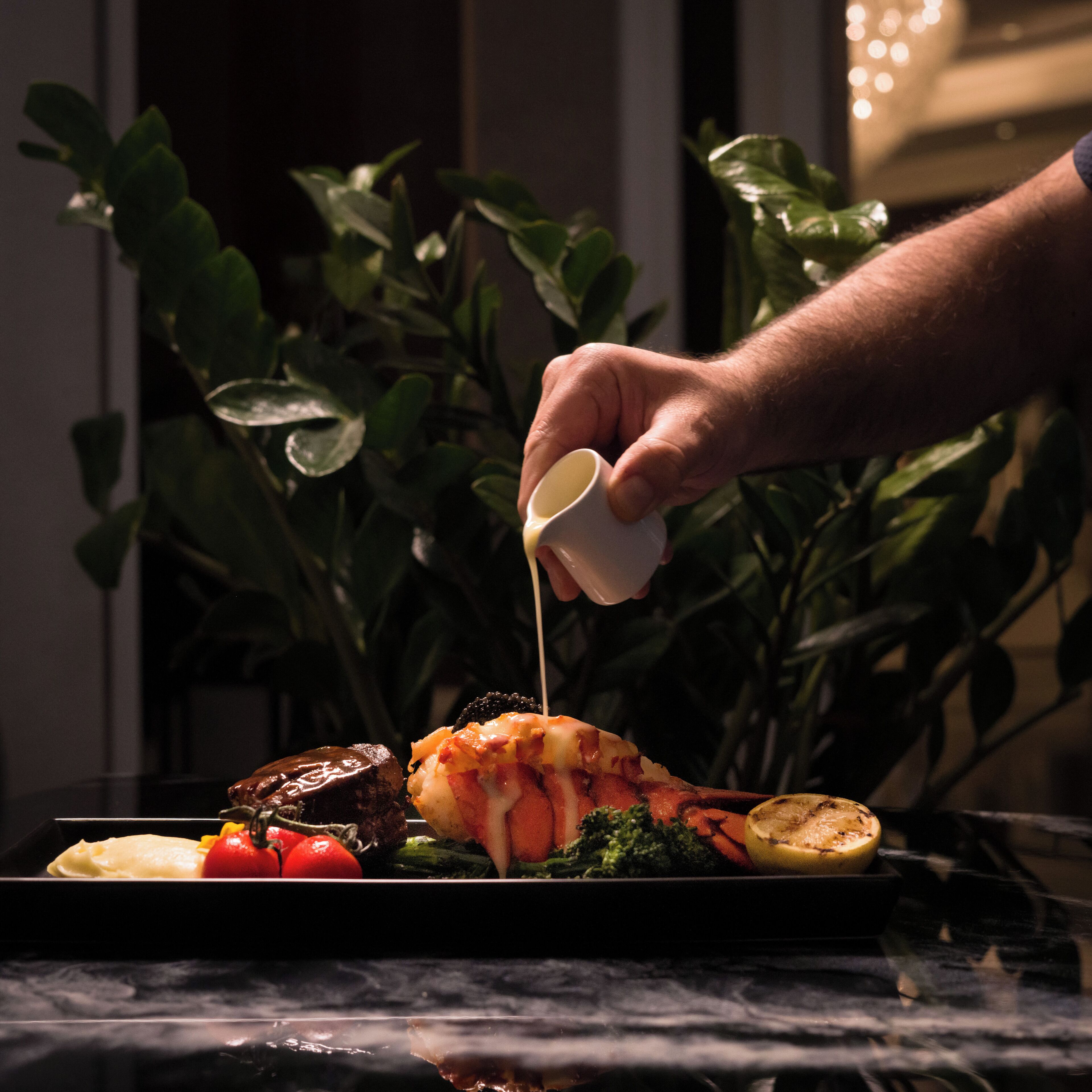 Una mano sirviendo salsa sobre una cola de langosta en un plato con vegetales.