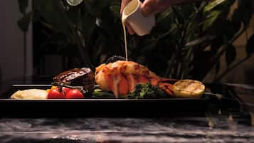 Una mano sirviendo salsa sobre una cola de langosta en un plato con vegetales.