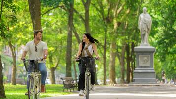 Two people riding bicycles on a park path with trees and a statue in the background.