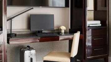 A hotel room with a wooden desk, a computer, a chair, a suitcase, and a TV mounted on the wall.