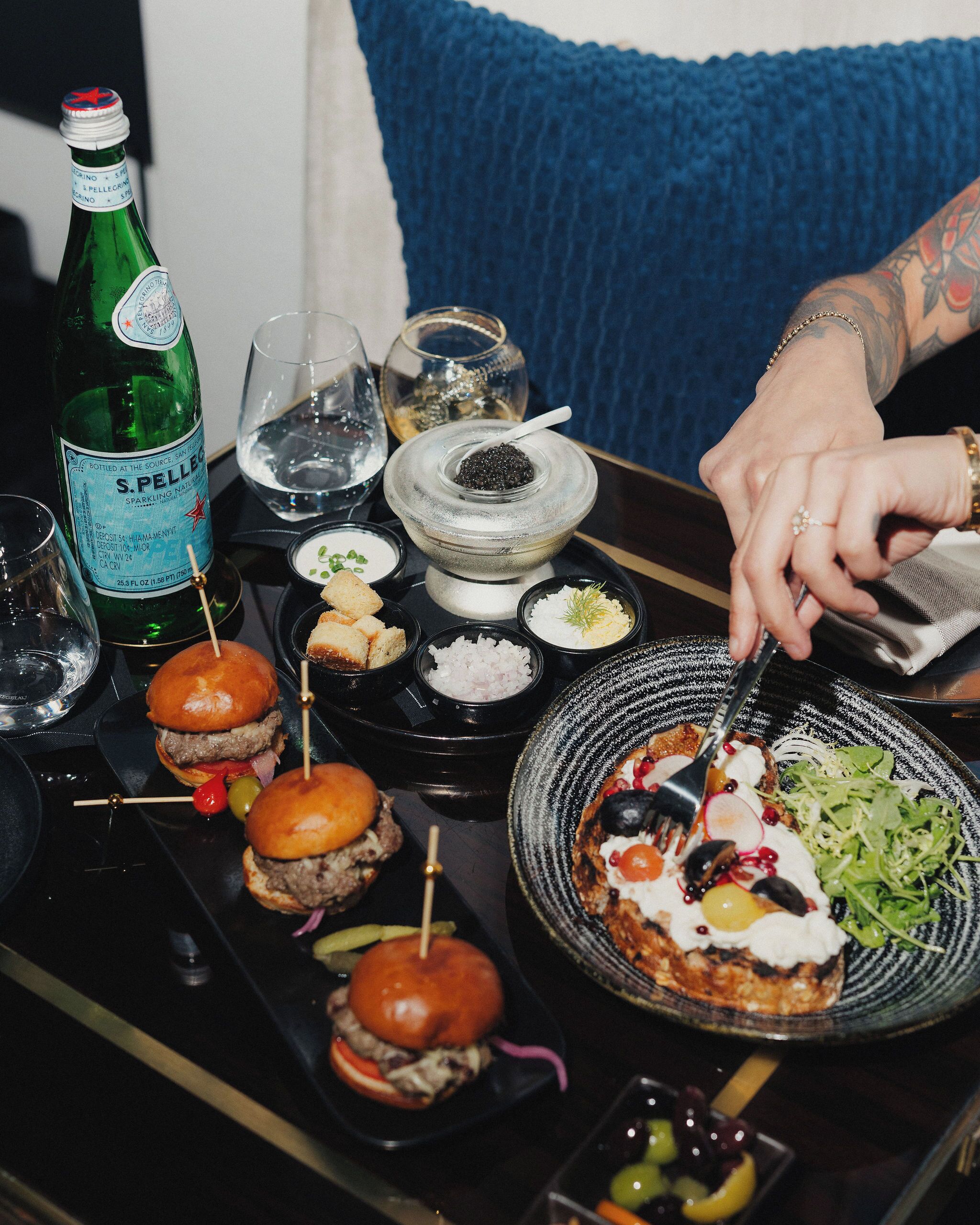 A dining table with food, a bottle of sparkling water, and a person cutting a dish.