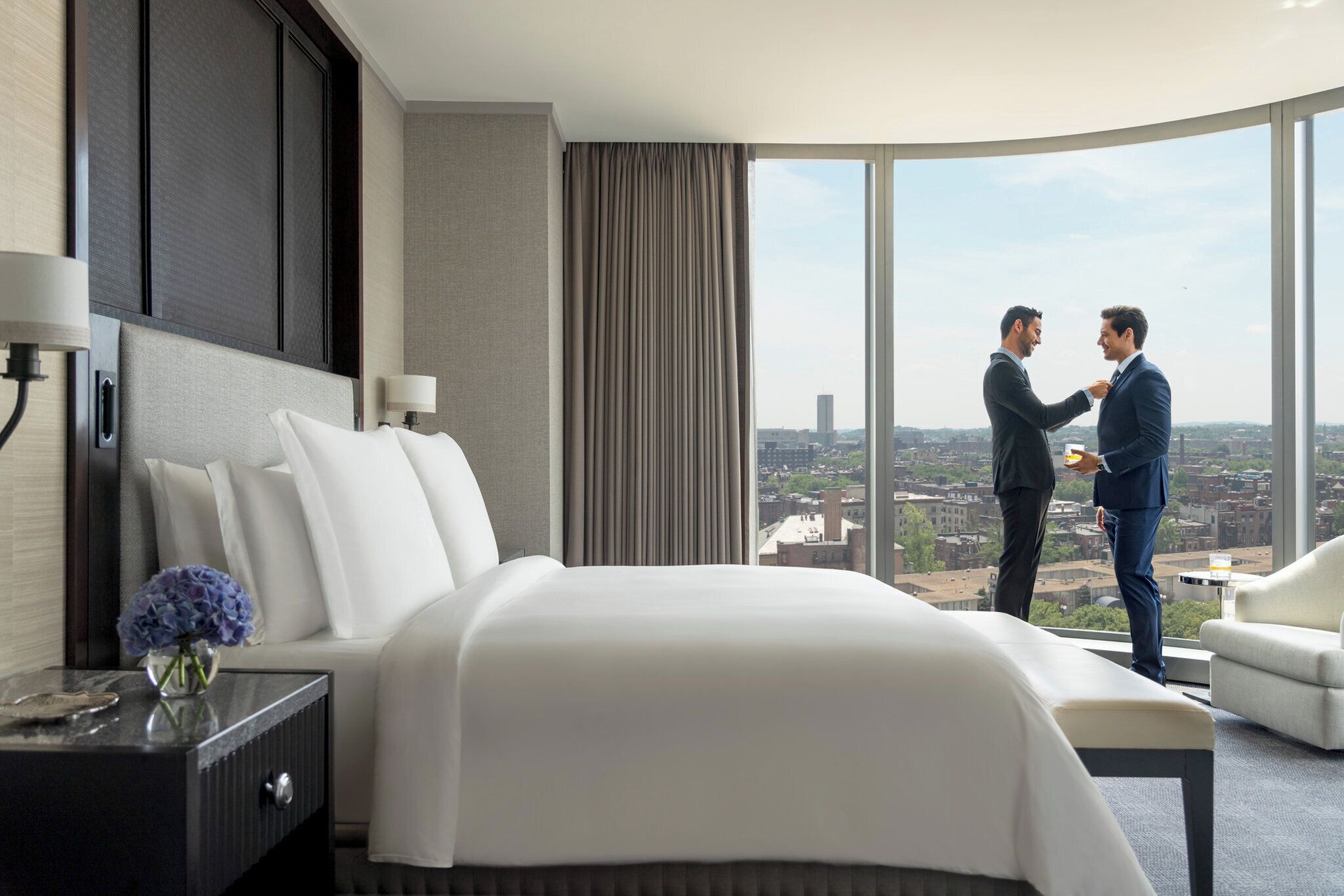 A hotel room with a large bed, a nightstand, a lamp, and a view of the city through large windows.