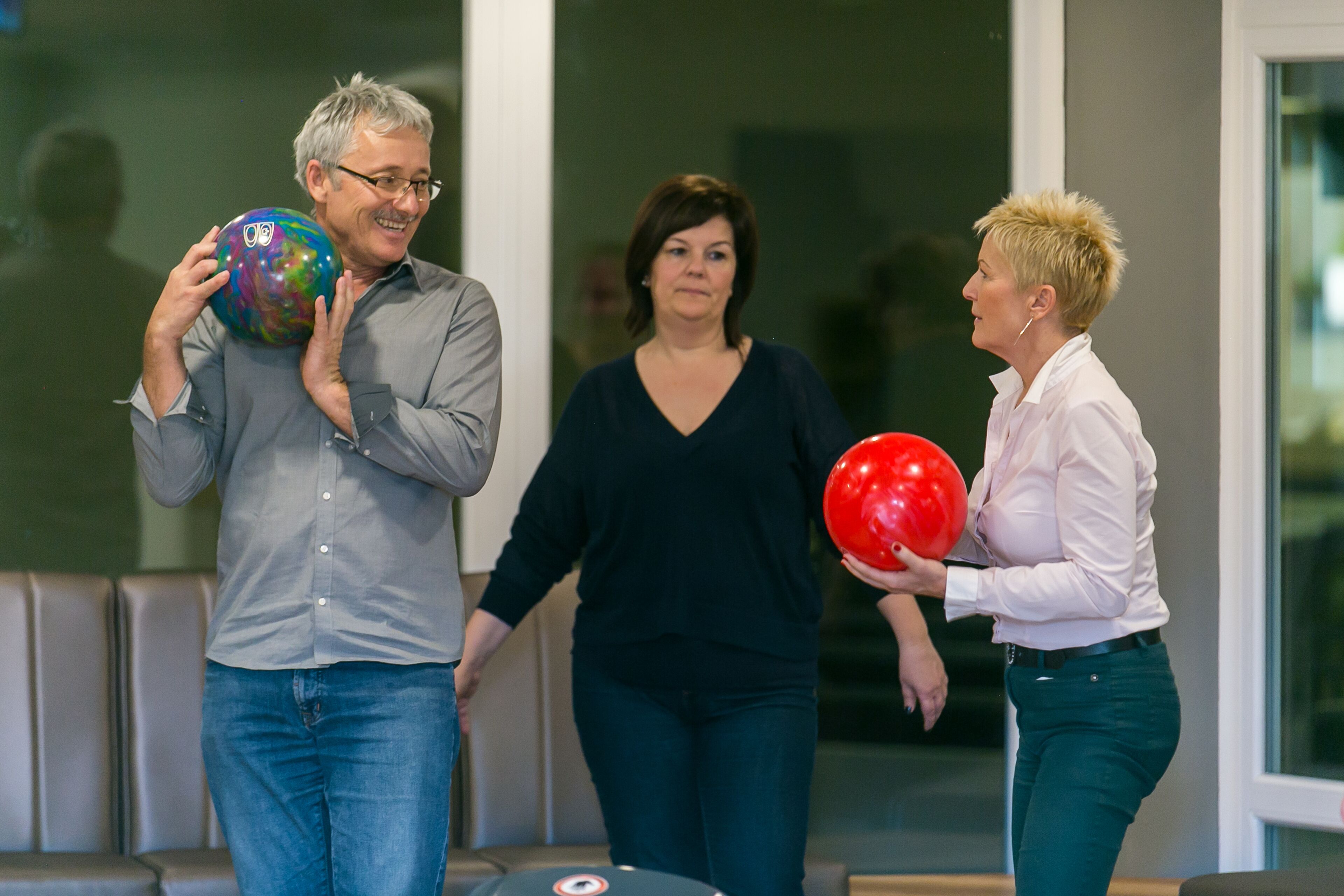 Trois personnes tenant des boules de bowling dans une pièce dotée d’une grande fenêtre et d’une piste de bowling.