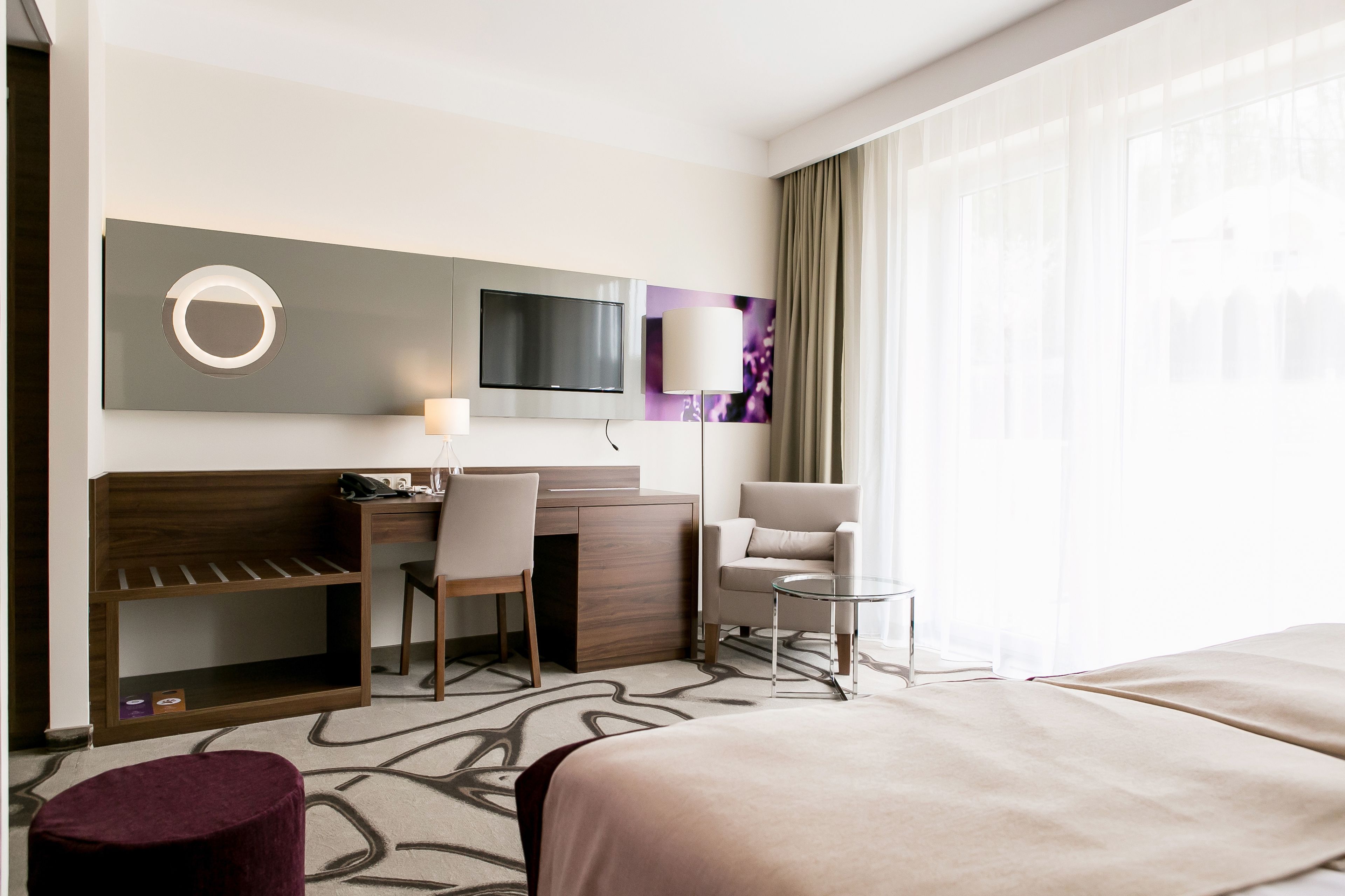 A hotel room with a bed, a desk with a chair, a TV mounted on the wall, a small round table, and a window with curtains.