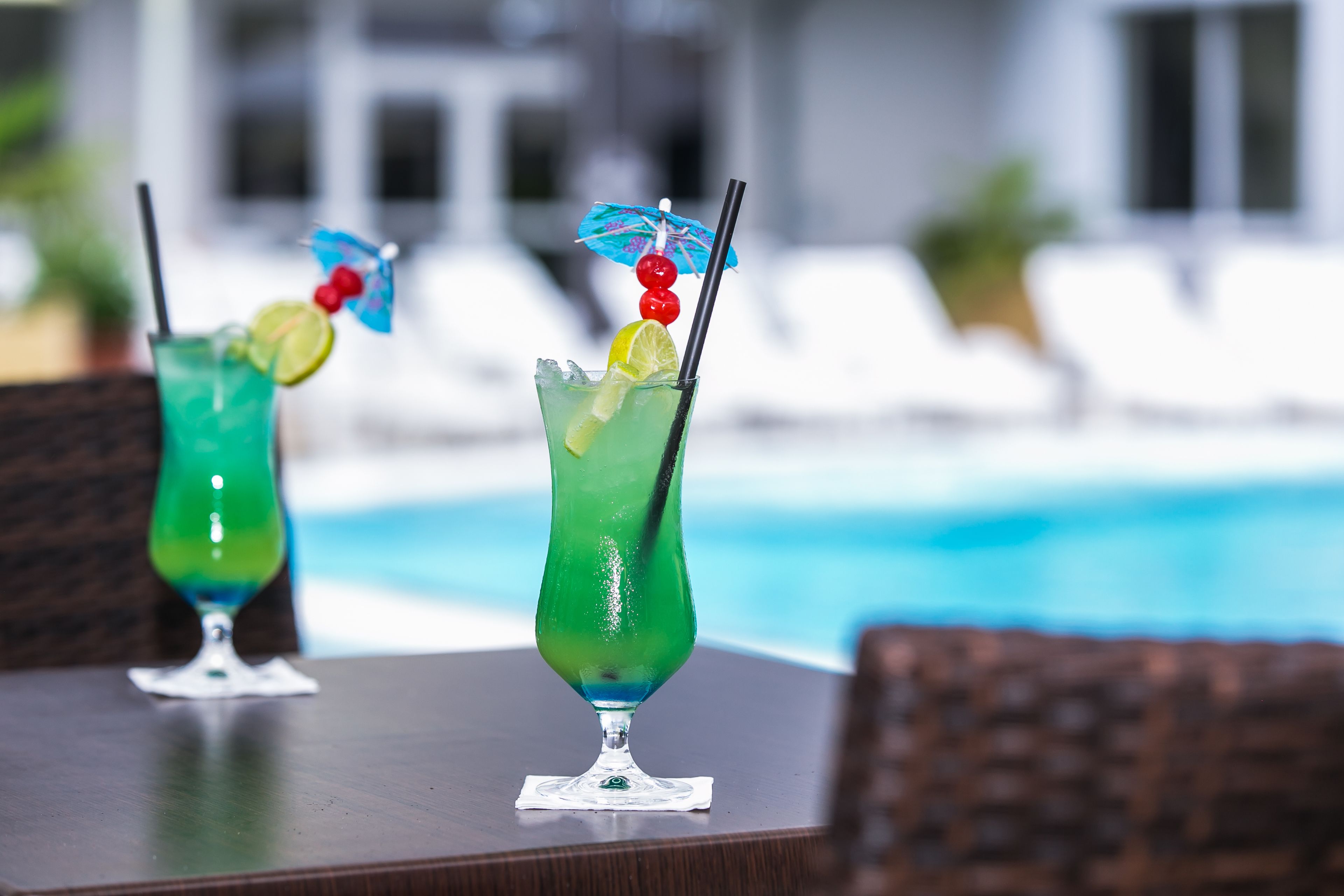 Two cocktails on a table by a poolside with lounge chairs in the background.