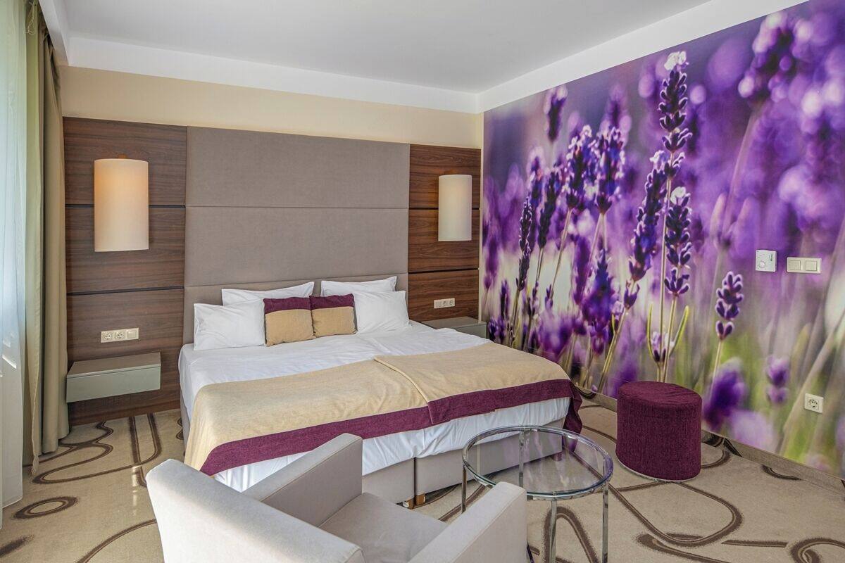 Quarto de hotel com uma cama grande, mesa de cabeceira, uma cadeira, mesa de vidro e uma parede com um mural floral.