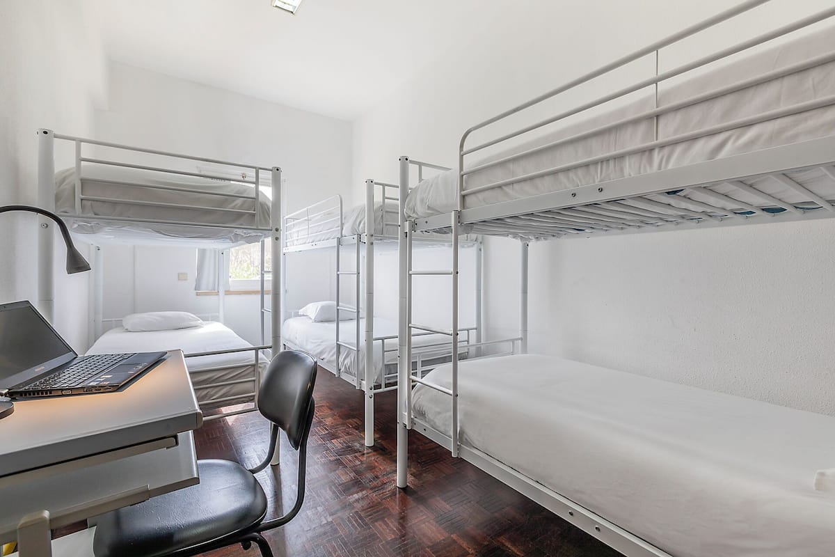 A dormitory room with bunk beds, a desk with a laptop, a chair, and a window.