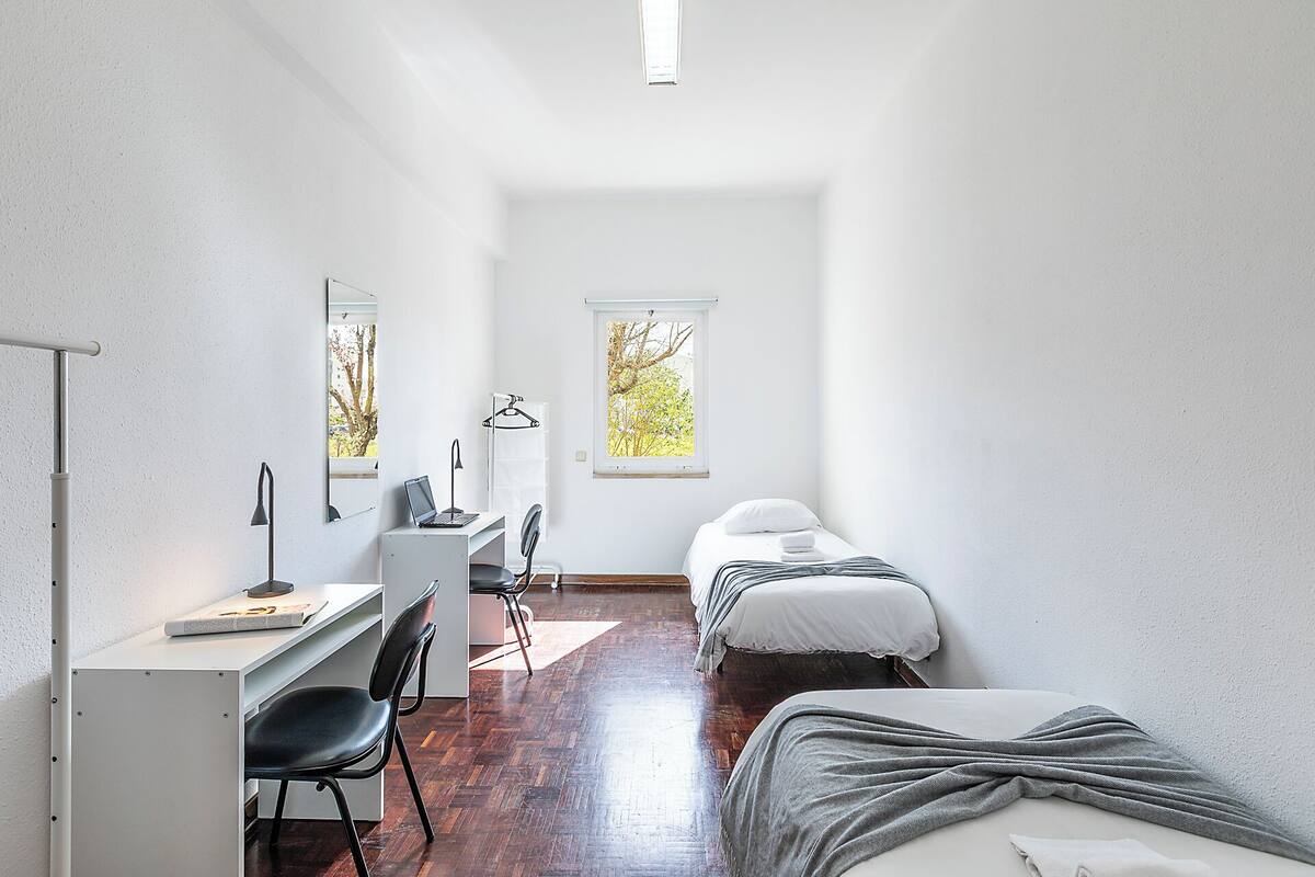 A compact room with a desk, two beds, a chair, and a window with a view of trees.