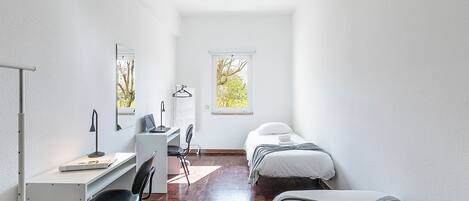 A compact room with a desk, two beds, a chair, and a window with a view of trees.