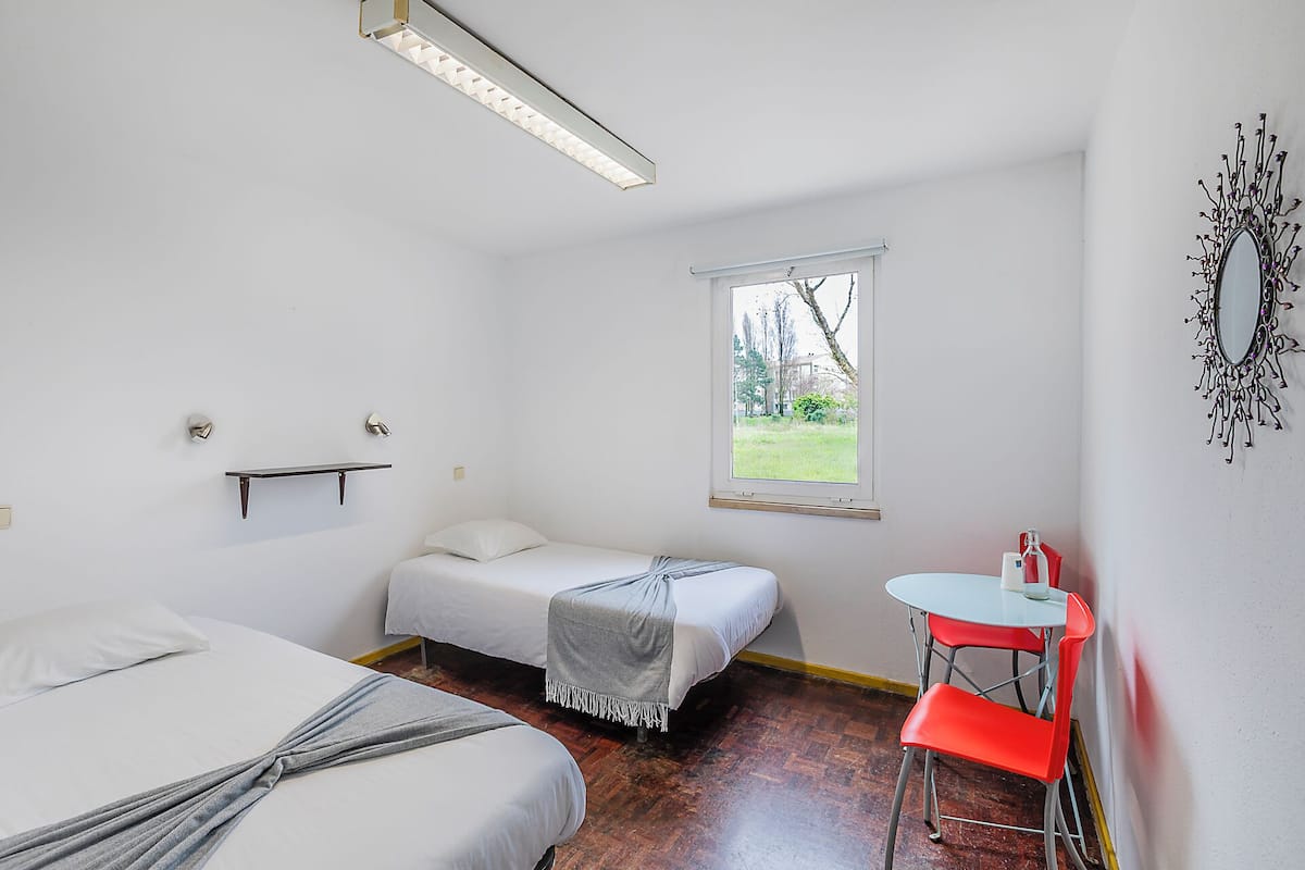 A room with two beds, a window with a view of greenery, a small table with a chair, and a wall-mounted mirror.