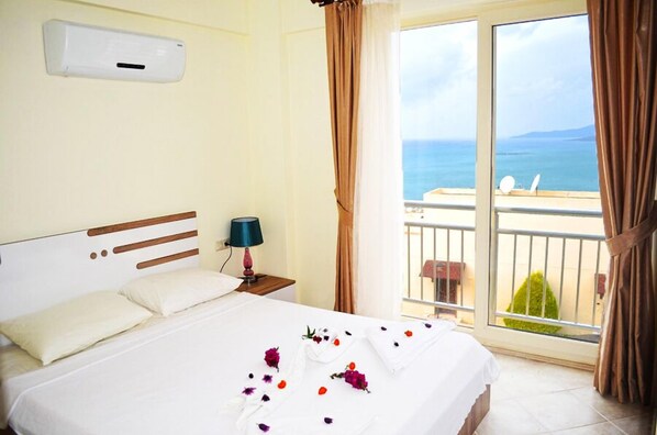 Habitación de hotel con una cama grande, una mesita de noche con lámpara, una ventana con vista al mar y un balcón con sillas.