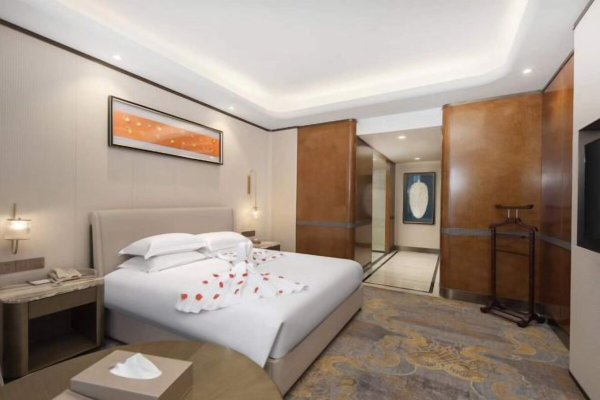A hotel room with a large bed, a bedside table with a lamp, a wall-mounted phone, and a framed picture above the bed.