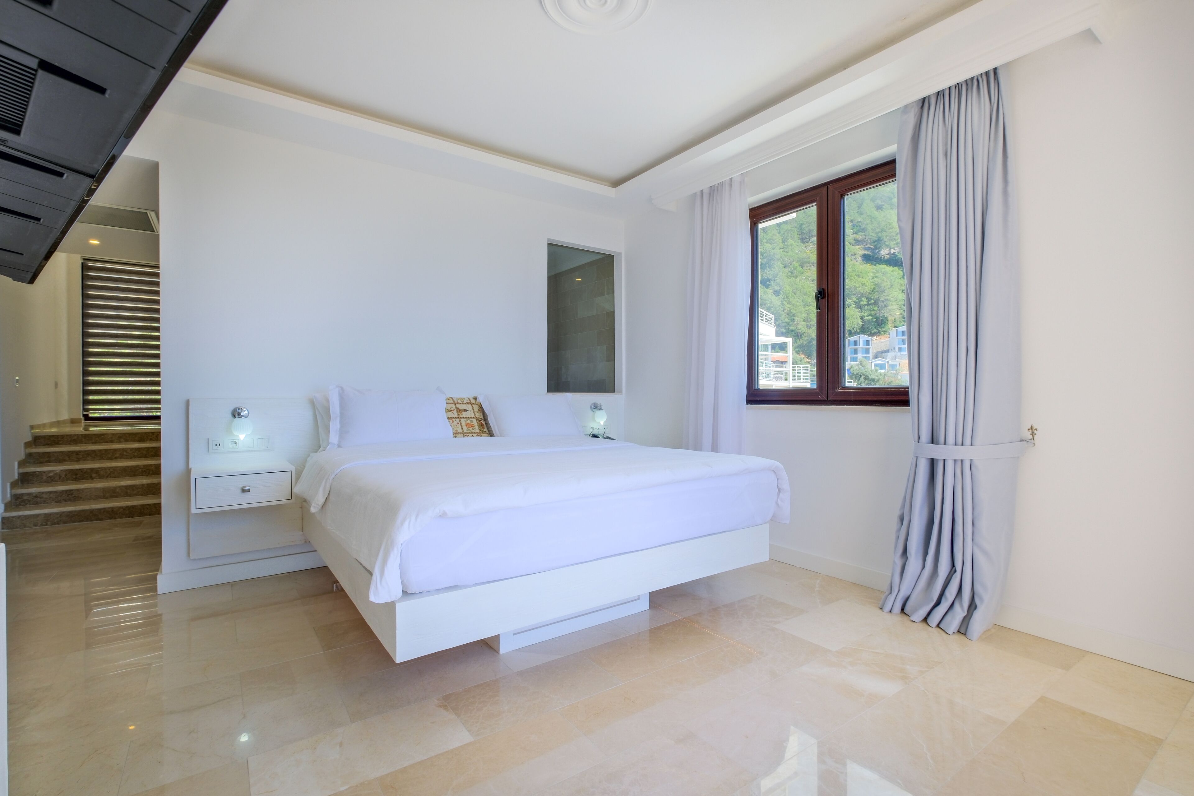A modern bedroom with a large bed, a nightstand, a window with curtains, and a staircase leading to another level.