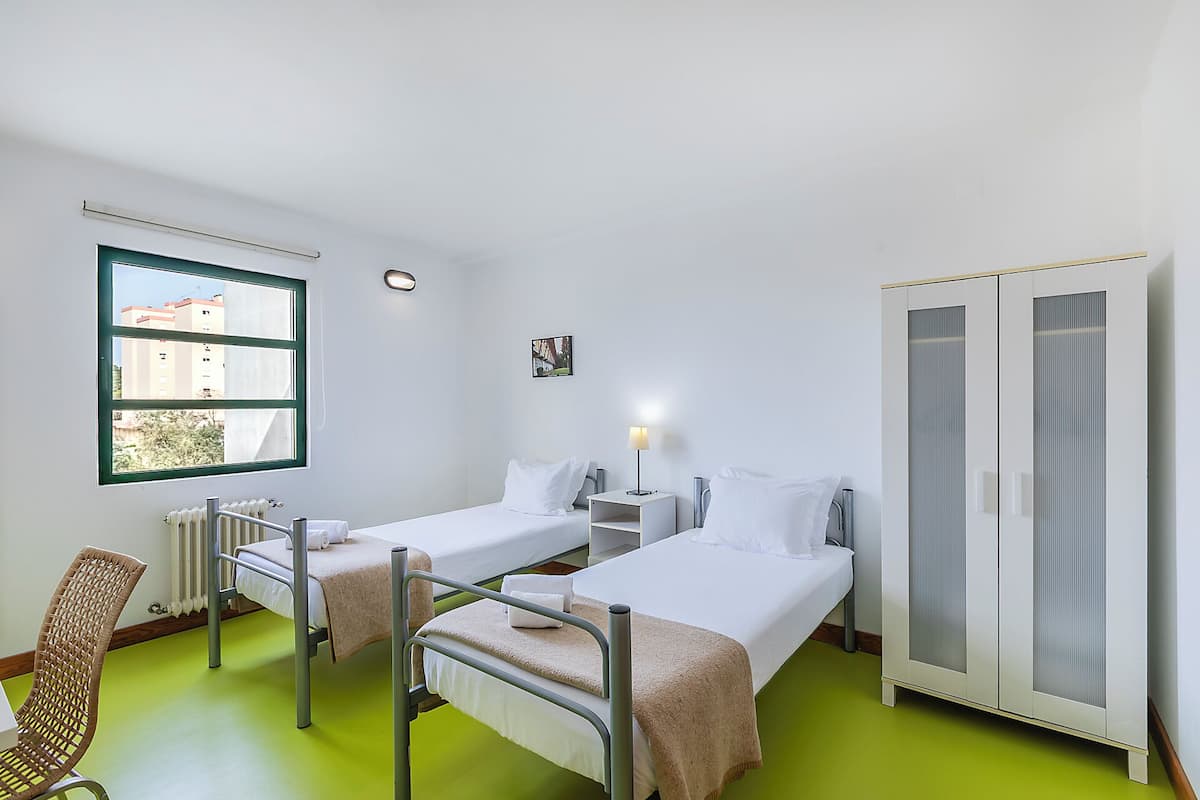 Een kamer met twee bedden, een stoel, een kledingkast, een radiator en een raam met uitzicht op gebouwen.