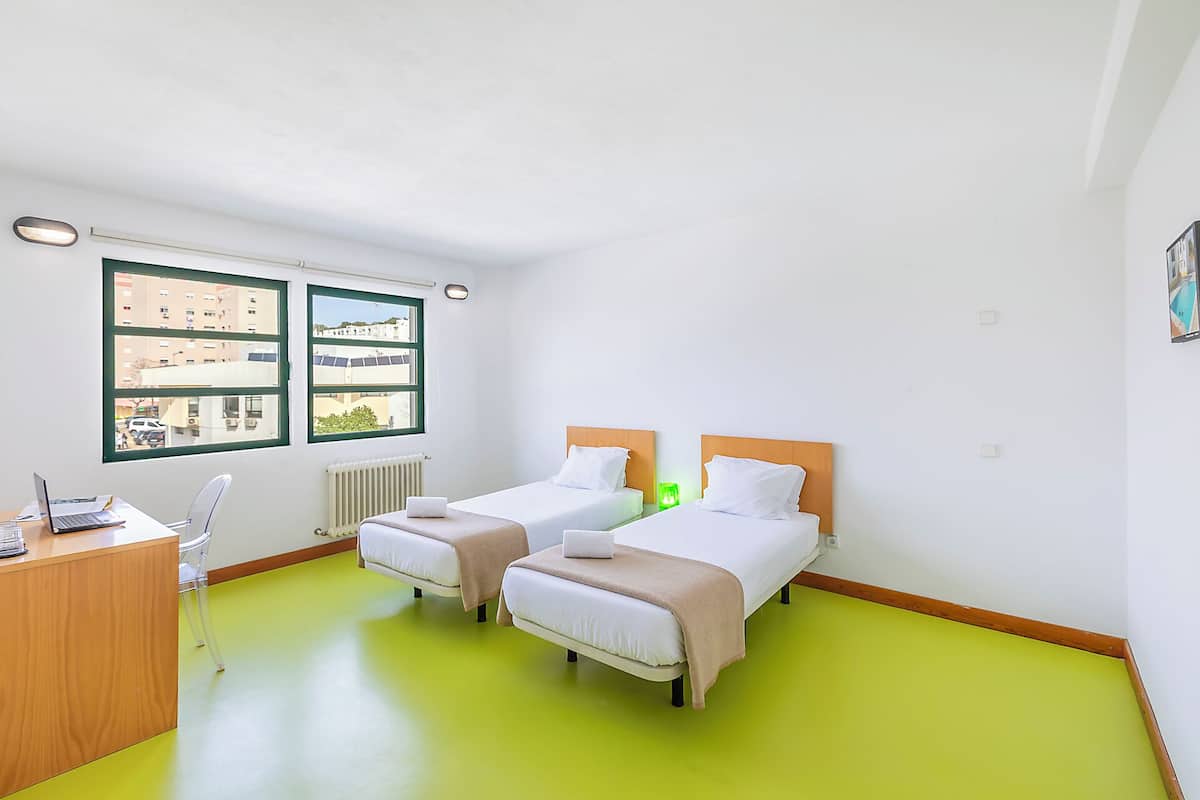 Hotelkamer met twee bedden, een bureau, een stoel, een radiator en een raam met uitzicht op gebouwen.