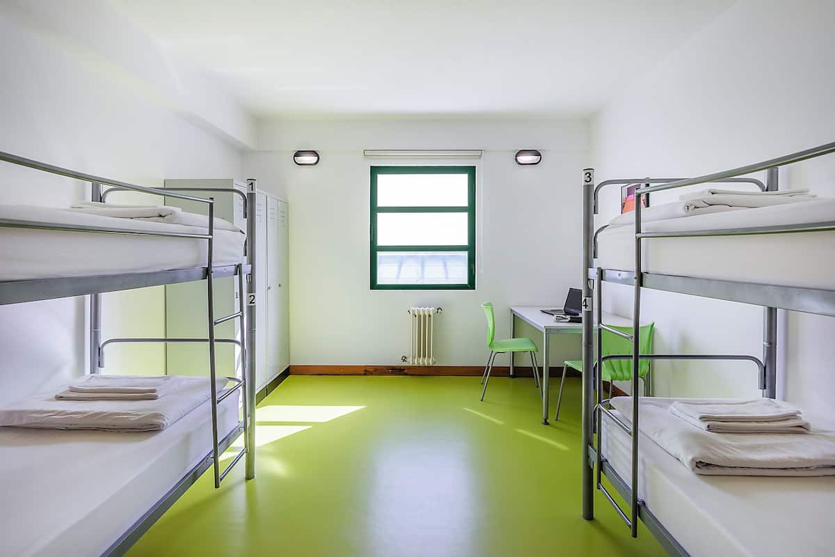 Een slaapzaal met stapelbedden, een radiator, een raam en een bureau met stoel.