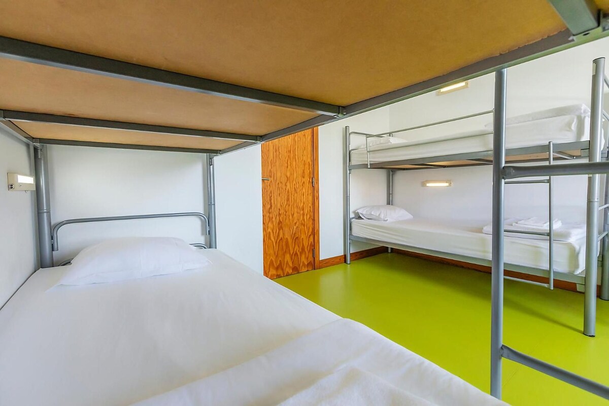 Quarto com beliche, uma cama de solteiro e dois beliches, painéis de madeira em uma parede e piso verde.