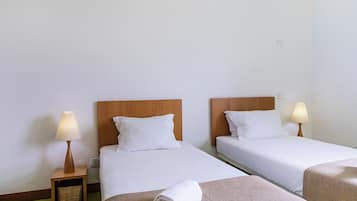 A hotel room with two beds, each with a wooden headboard and white bedding. There are bedside lamps on wooden nightstands, and a rolled towel on the floor.