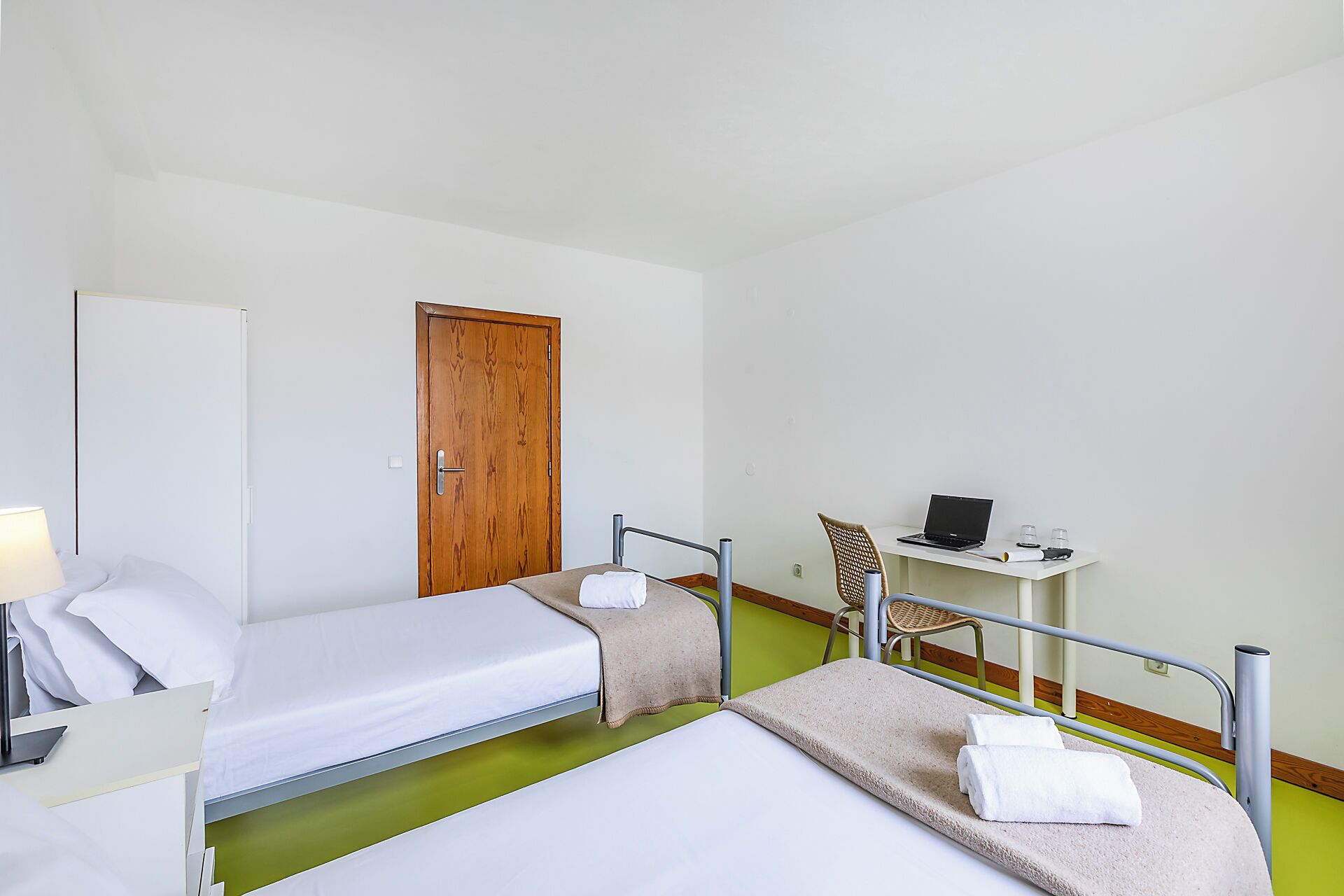 A hotel room with two single beds, a desk with a laptop, a chair, and a wooden door.