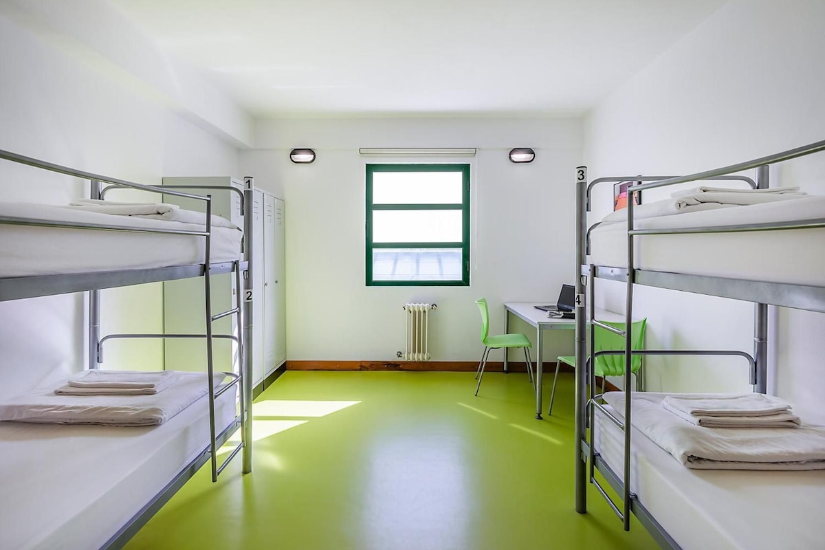 A dormitory room with bunk beds, a desk, a chair, and a radiator.