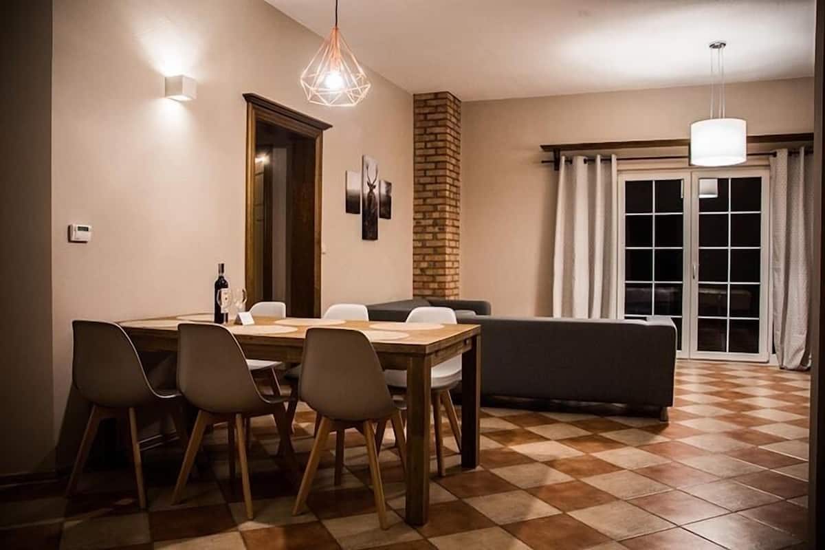 A dining area with a wooden table and chairs, a brick accent wall, and a large window with curtains.