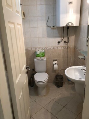 Ein kleines Badezimmer mit Toilette, Waschbecken und Dusche. Toilettenpapier und ein Mülleimer sind zu sehen.