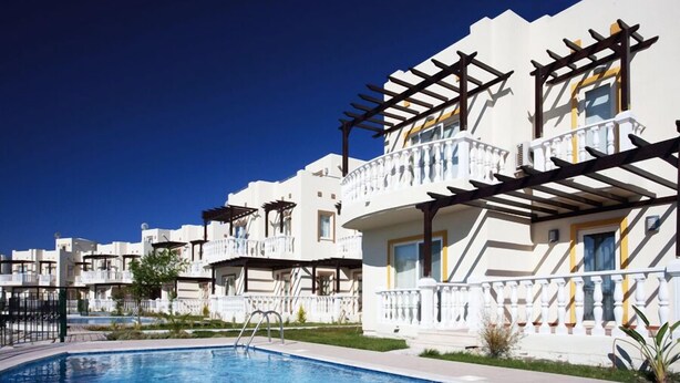 Um resort com piscina, edifícios brancos com varandas e um céu azul límpido.