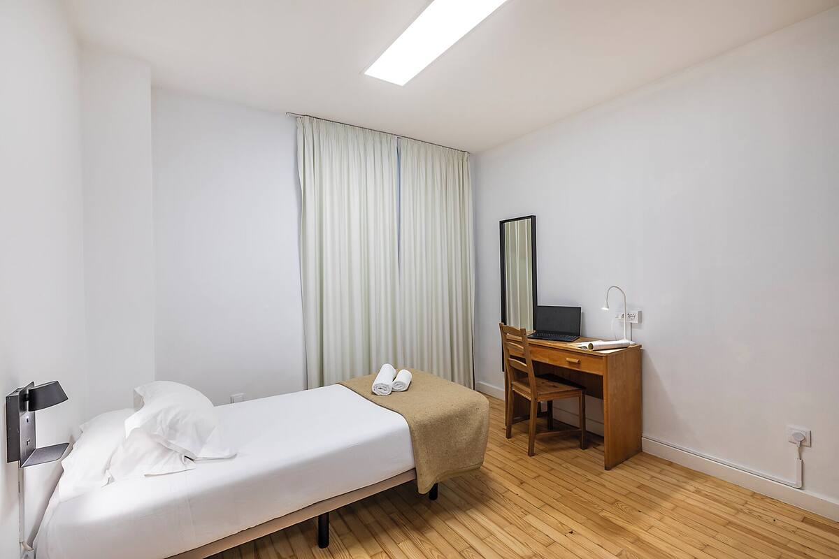 Une chambre à coucher avec un lit simple, un bureau en bois, une chaise, un miroir et une table de chevet.