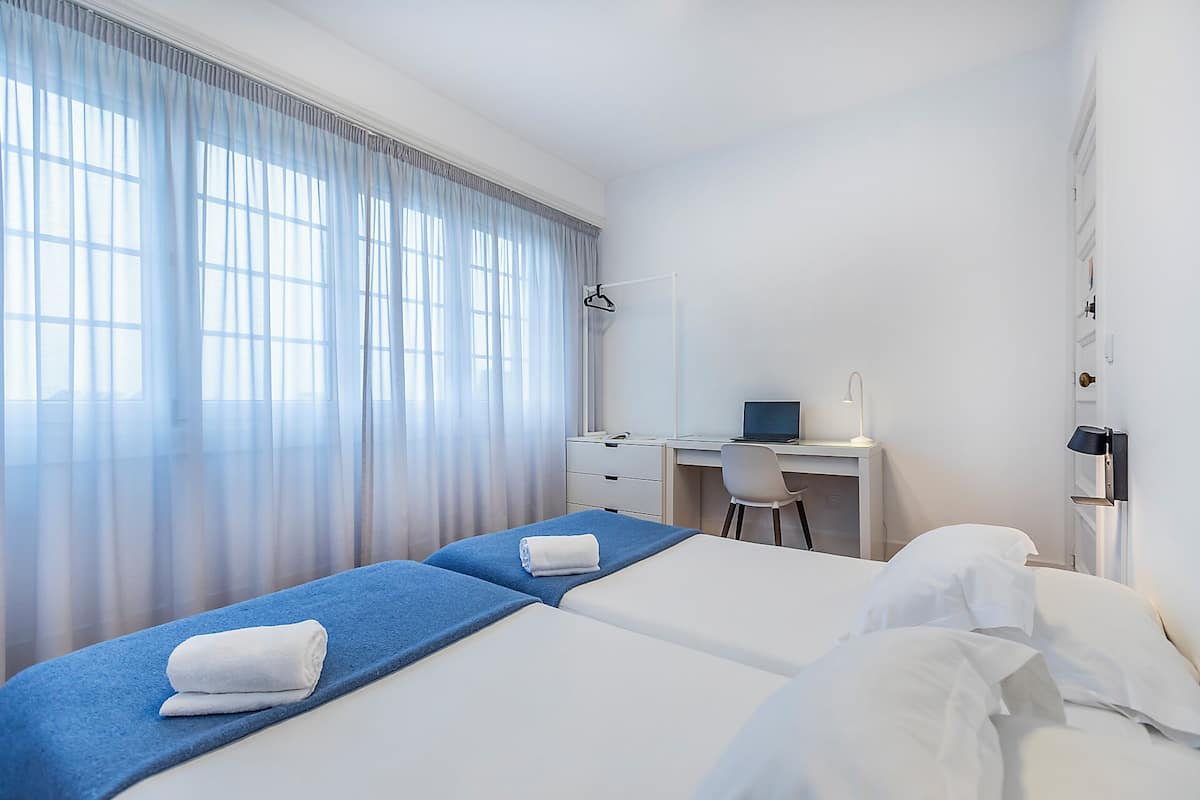 Une chambre d’hôtel avec deux lits, un bureau, une chaise et une grande fenêtre avec des rideaux légers.