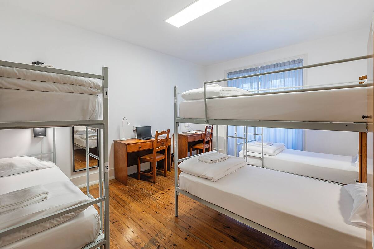 Une chambre avec un lit superposé, un bureau et une chaise, une fenêtre avec des rideaux et un plancher en bois.