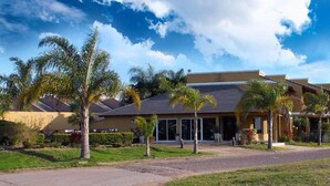 A hotel with a modern design, featuring a flat roof and large windows. There are palm trees and a well-maintained lawn in front.