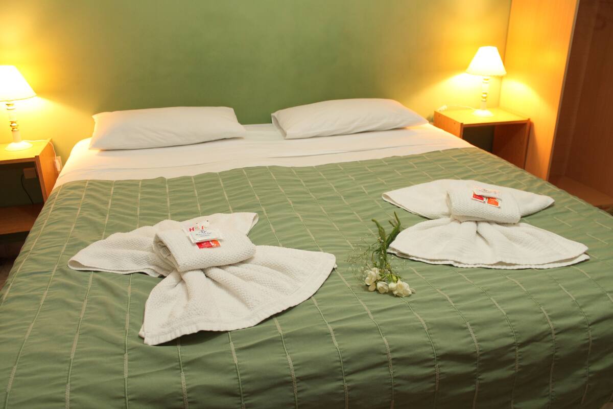 Una cama bien hecha con dos toallas dobladas en forma de corazón, una colcha verde y dos lámparas de noche.
