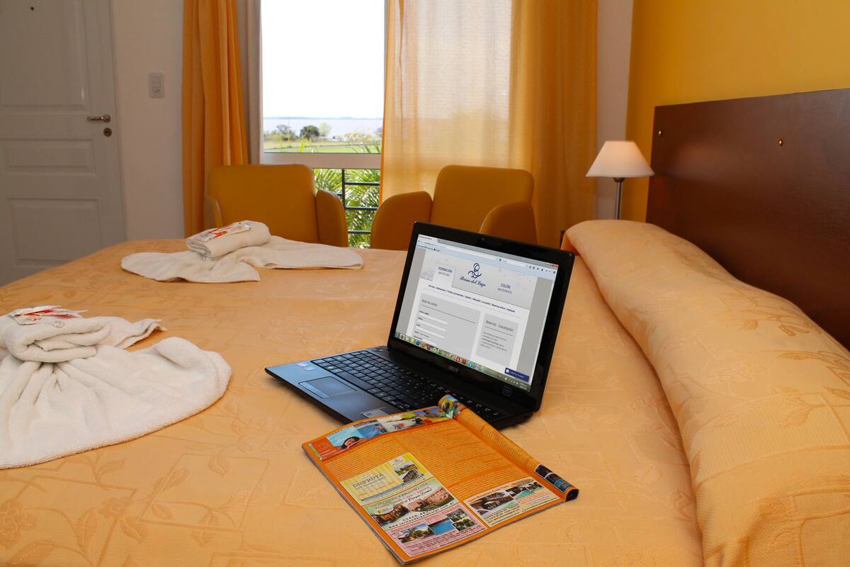 Habitación de hotel con una cama, una computadora portátil, una revista y una ventana con cortinas.
