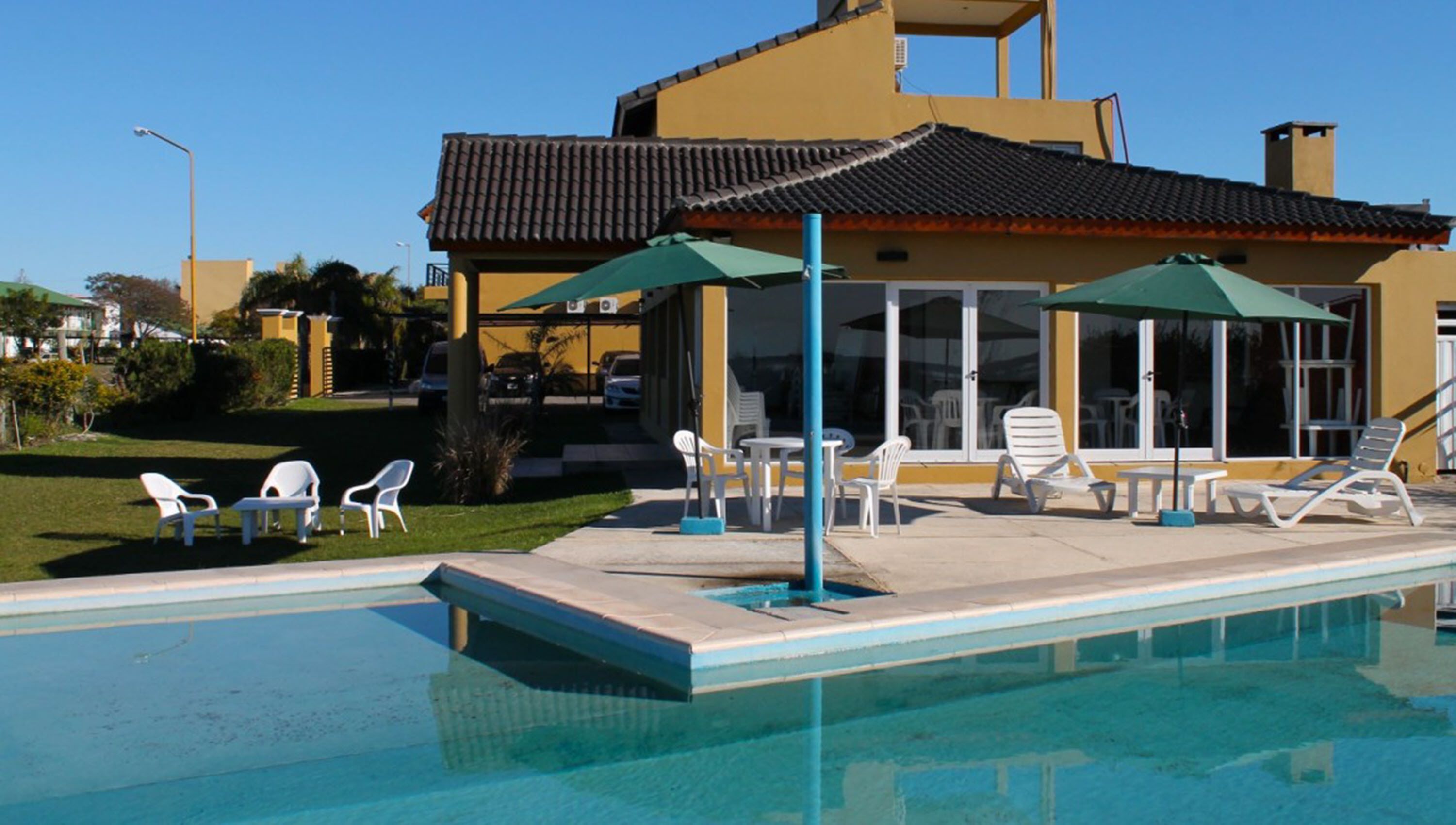 A swimming pool with lounge chairs and umbrellas, a yellow building with a tiled roof, and a clear blue sky.