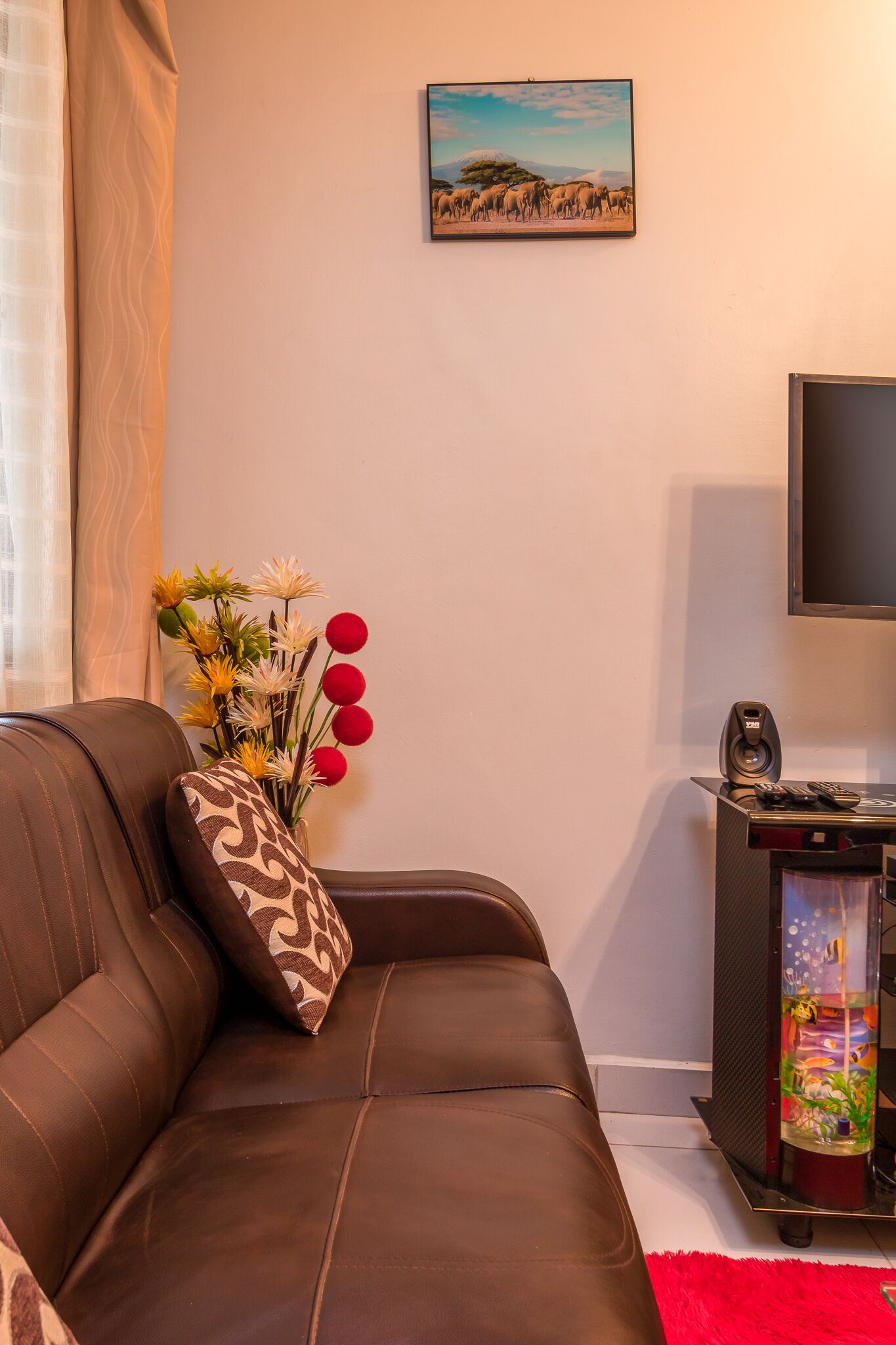 A brown leather sofa with a patterned pillow, a wall-mounted TV, a decorative vase with flowers, and a framed picture above the sofa.