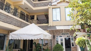 Un hotel de varios pisos con un balcón en el segundo piso, un área de estar exterior cubierta con mobiliario de mimbre y un mostrador de recepción.