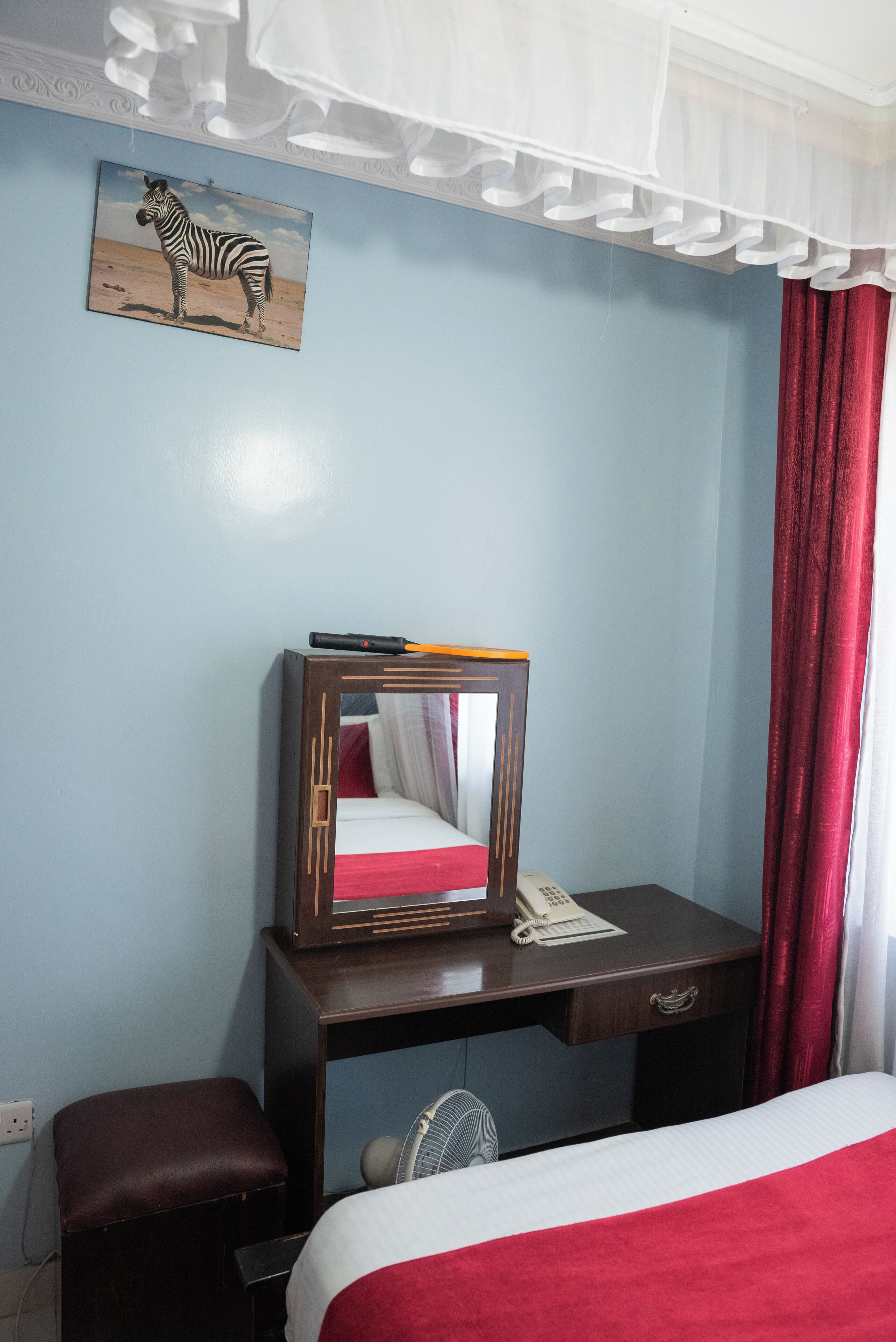 A hotel room with a bed, a desk with a telephone and papers, a fan, a zebra painting, and a red curtain.