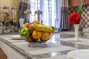 A kitchen counter with a fruit bowl containing apples, bananas, and oranges, a white vase with a red flower, and a checkered backsplash. - Javilla Eagles Safari Guest House (Nairobi)