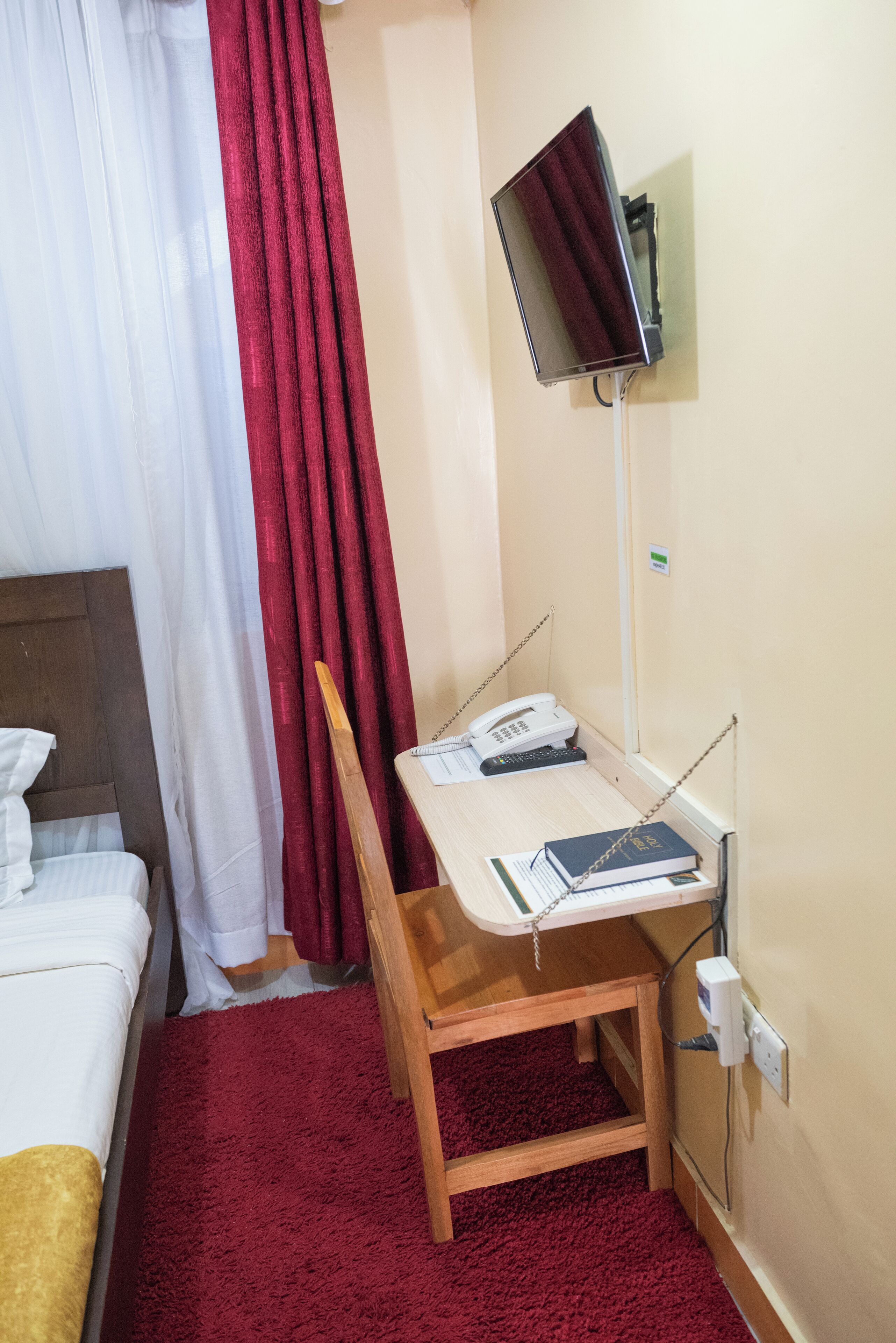 A hotel room with a bed, a desk with a telephone and a book, a chair, and a TV mounted on the wall.
