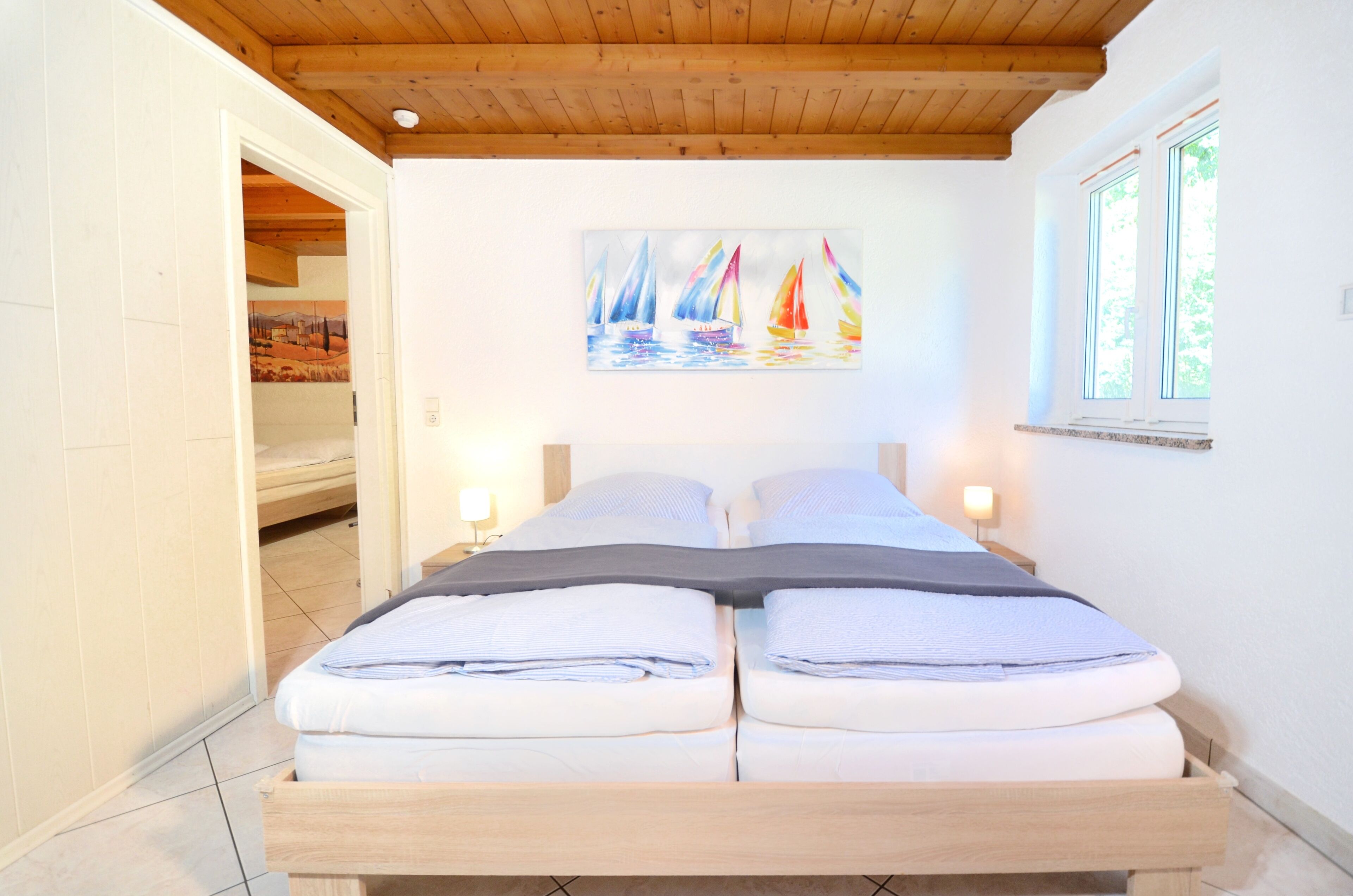 Ein Doppelbett mit zwei Kissen, ein Segelbootgemälde an der Wand, ein Fenster mit Aussicht und eine Holzbalkendecke.