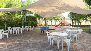 Un'area patio esterna con tavoli e sedie di plastica bianca, un'area giochi e un'ampia tettoia che offre ombra.